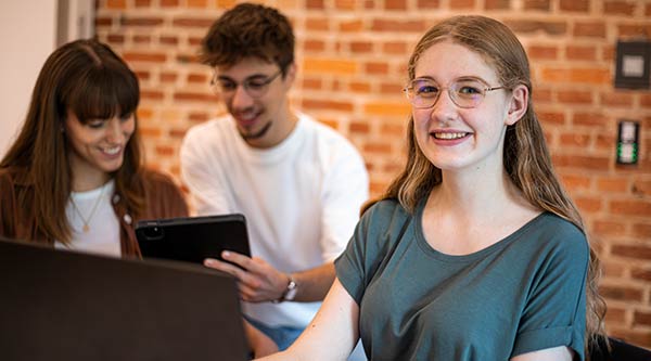 Eine Jugendliche sitzt lächelnd vor einem Laptop und zwei weitere blicken im Hintergrund auf ein Tablet.