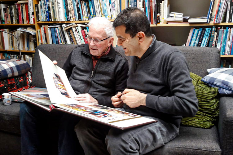 Zwei Männer sitzen auf einem Sofa vor einem Bücherregal und schauen sich ein Fotoalbum an