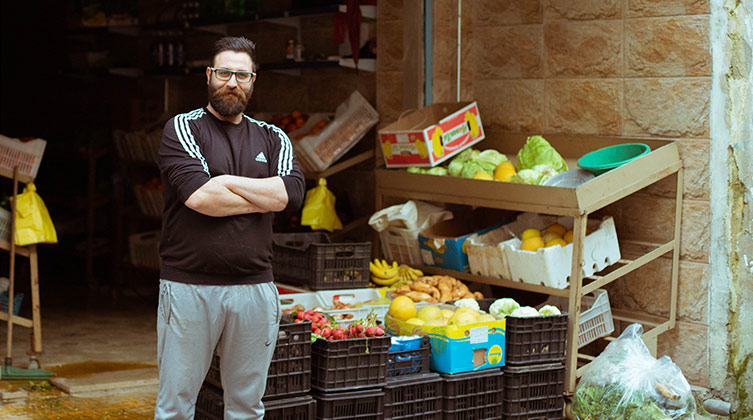 Ein Mann steht mit verschränkten Armen vor mehreren Regalen mit Obst und Gemüse