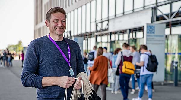 Lächelnder Mann mit Open Doors Lanyard um den Hals und Halle von außen mit Menschen im Hintergrund