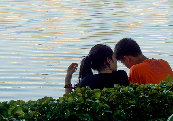 Symbolbild: Ein junges Paar sitzt hinter einer grünen Hecke am stillen Gewässer