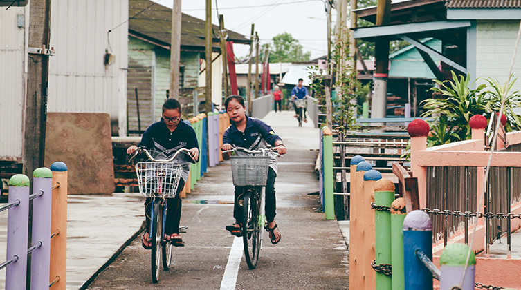 Zwei malaysische Mädchen fahren auf dem Fahrrad frontal auf die Kamera zu und hinter ihnen sieht man die von Häusern umgebene Gasse