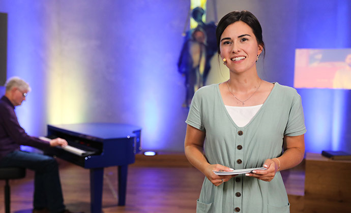 Eine junge Frau mit grünem Sommerkleid spricht zu dem Zuschauer und im Hintergrund sitzt ein älterer Mann an einem Flügel und spielt