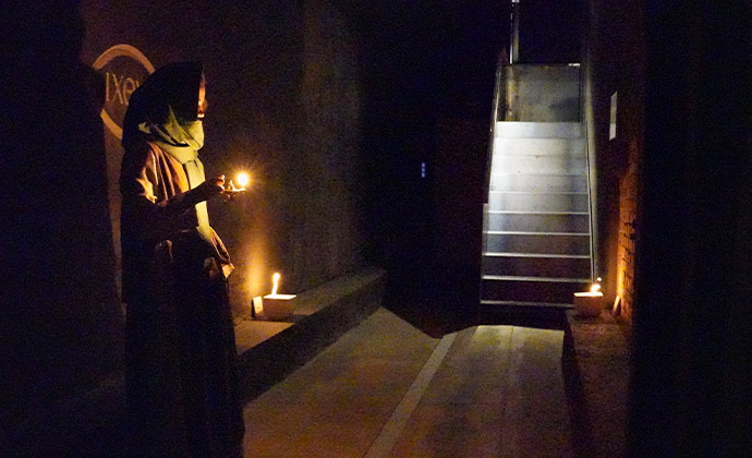 Ein sehr dunkler Raum in den hinten eine helle Treppe hineinführt und einer verschleierten Person am Rand, die ein Kerzenlicht hält