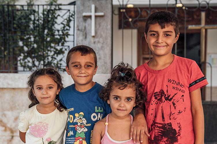 Vier kleine Kinder mit bunter Kleidung stehen eng nebeneinander vor einem Kirchengebäude