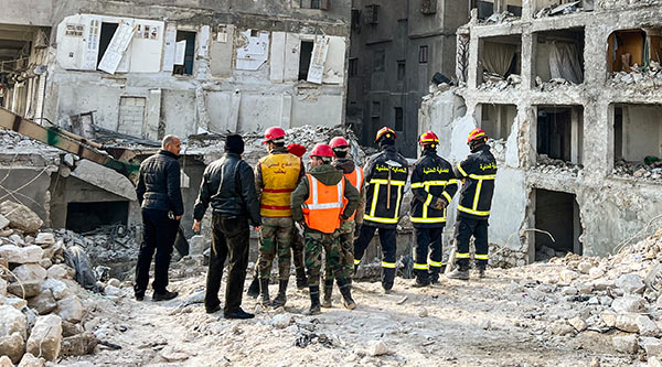 Eine Gruppe Sicherheitskräfte, Helfer und Bauarbeiter stehen vor dem Krater nach dem Erdbeben, kaputte Häuser im Hintergrund