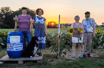 Ein älteres Ehepaar, deren Tochter und Enkelin und ein Hund stehen vor einem Sonnenblumenfeld, links und rechts eine Kassentonne mit Aufschrift