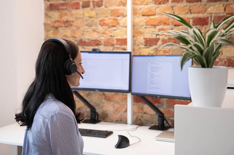 Eine lächelnde Frau mit schwarzen Haaren sitzt mit Headset auf dem Kopf vor zwei Bildschirmen und einer Ziegelwand am Schreibtisch