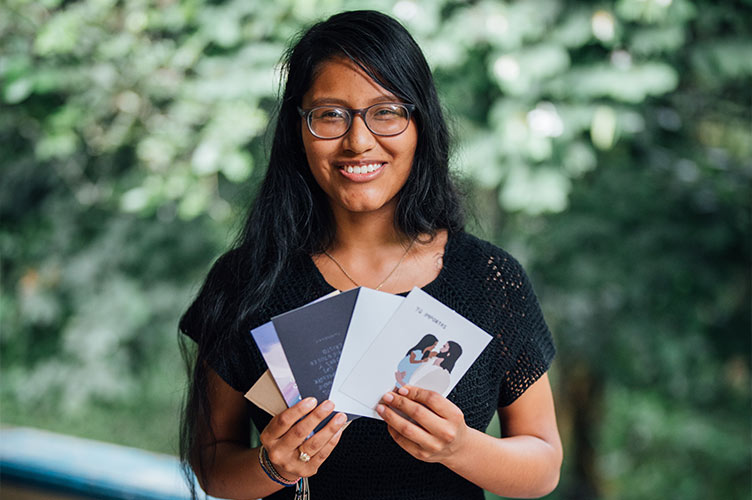 Eine südamerikanische junge Frau hält lächelnd Ermutigungskarten in der Hand und steht vor grünen Büschen