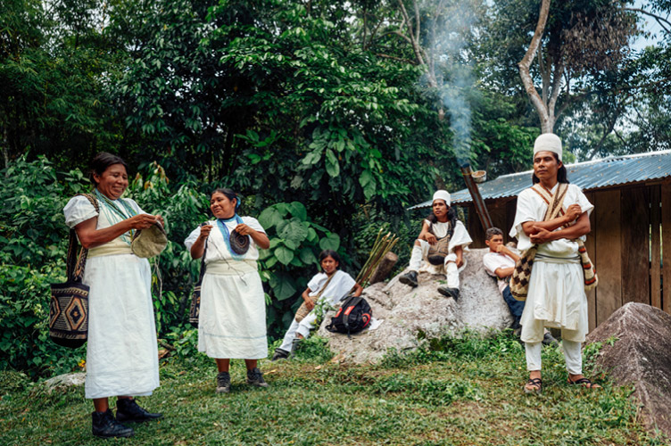 Eine Gruppe der indigenen kolumbischen Bevölkerung in weißer Tracht steht vor einem Wald und einer Hütte, dazwischen die Überschrift des Open Doors Jugendtags 2024 zu Kolumbien