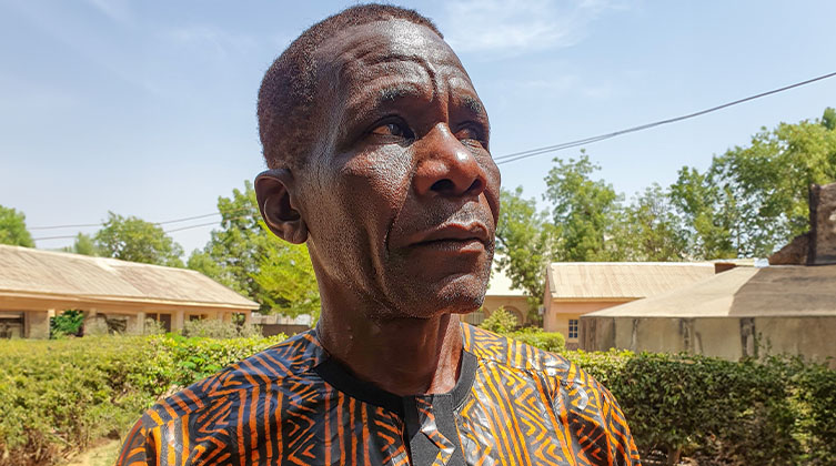 Ein Mann aus Nigeria steht in seinem Dorf vor grünen Hecken. Er wird im Portrait abgebildet. Im Hintergrund sind einzelne kleine Häuser und einige Bäume zu sehen.