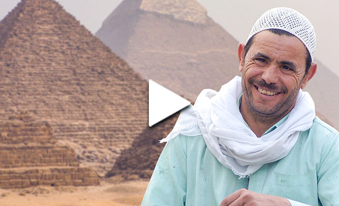 Ein Mann steht vor den Pyramiden in Gizeh. Er trägt ein türkisfarbenes Gewand und ein weißes Halstuch und eine weiße Kopfbedeckung.