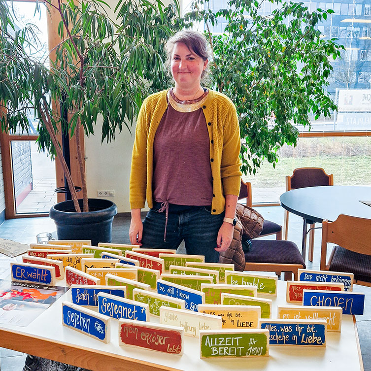 Eine Unterstützerin steht vor ihrem Stand. Sie verkauft bunt bemalte Holztafeln und unterstützt mit dem Erlös ihre verfolgten Glaubensgeschwister.