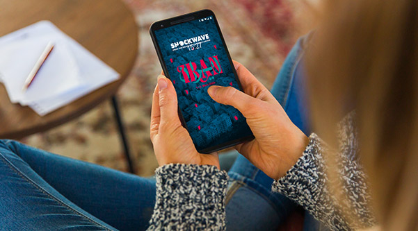 Eine junge Frau sitzt im Schneidersitz und hält in der Hand ein Handy mit dem Shockwave Screensafer