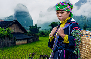 Eine Frau in Laos steht vor einer Hütte auf einer grünen Wiese. Im Hintergrund ist ein Gebirge.