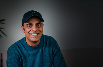 Ein Mann mit blauem Langarmshirt und Kappe vor einem neutralen grauen Hintergrund