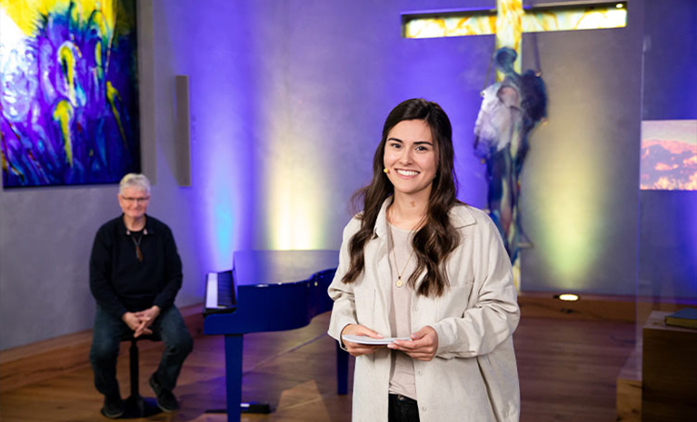Eine Frau mit Moderationskarten in der Hand. Im Hintergrund sitzt ein Mann am Klavier