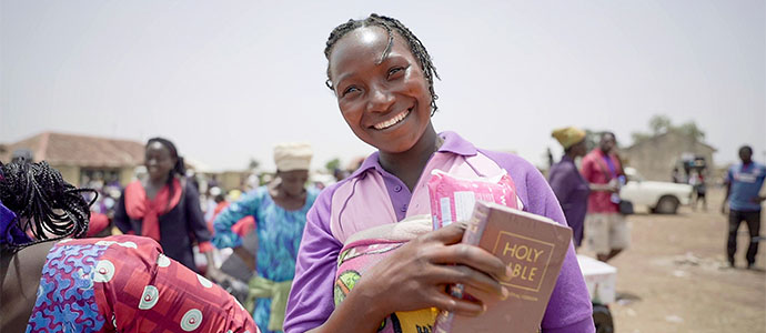 Eine afrikanische Frau steht mit anderen Menschen vor einem Dorf und lächelt mit einer Bibel in der Hand