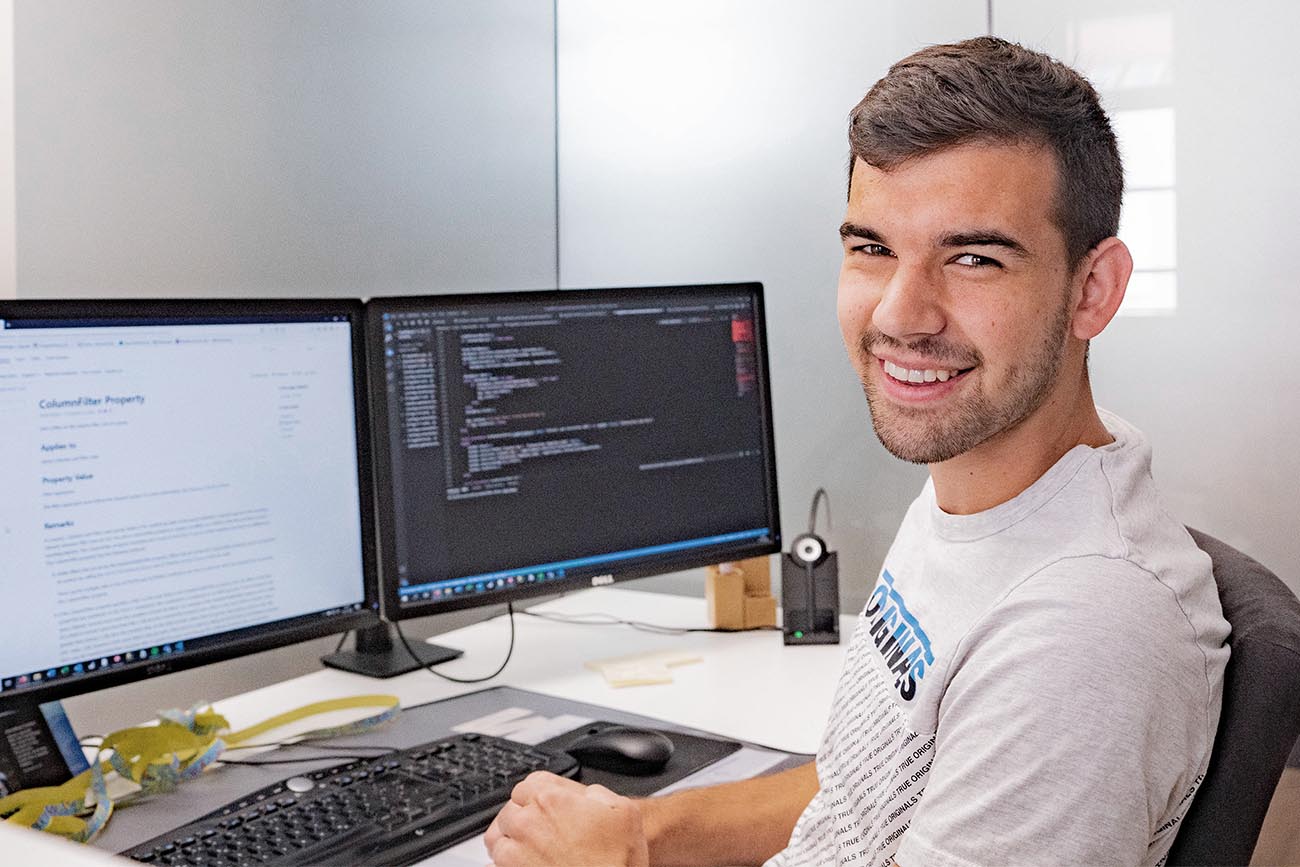 Ein junger Mann sitzt an einem Schreibtisch mit Computer und lächelt