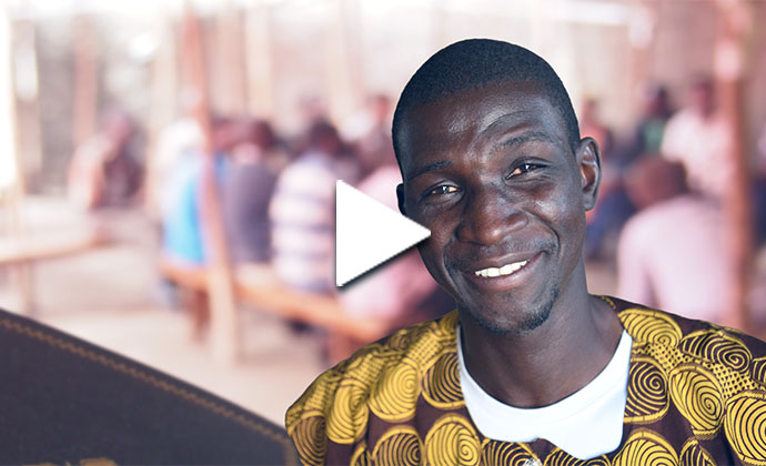 Ein afrikanischer Christ lächelt in Richtung des Betrachters. Im Hintergrund sitzen einige Menschen auf Bänken an Tischen