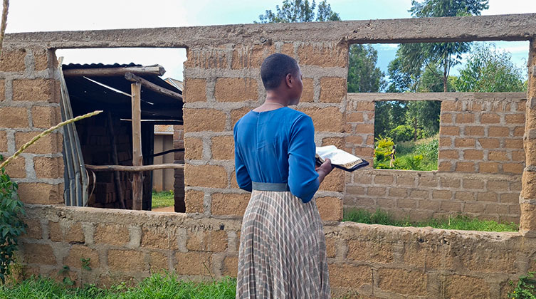 Eine afrikanische Frau mit einem blau-braunen Kleid steht vor einer einfachen Mauer aus Lehm und hält eine aufgeschlagene Bibel in der rechten Hand