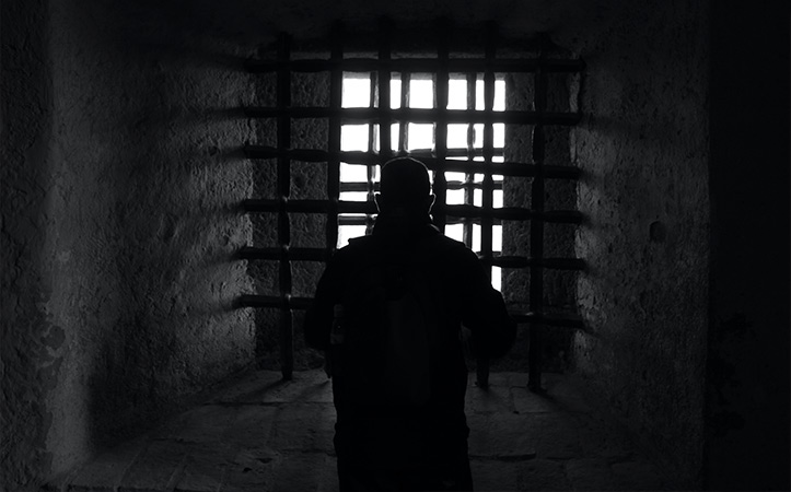 Mann steht in einem dunklen Raum vor Gittern hinter denen ein helles Licht scheint