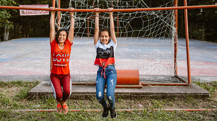 Zwei kolumbianische Mädchen hängen an einer Turnstange vor einem Sportplatz und lächeln in die Kamera