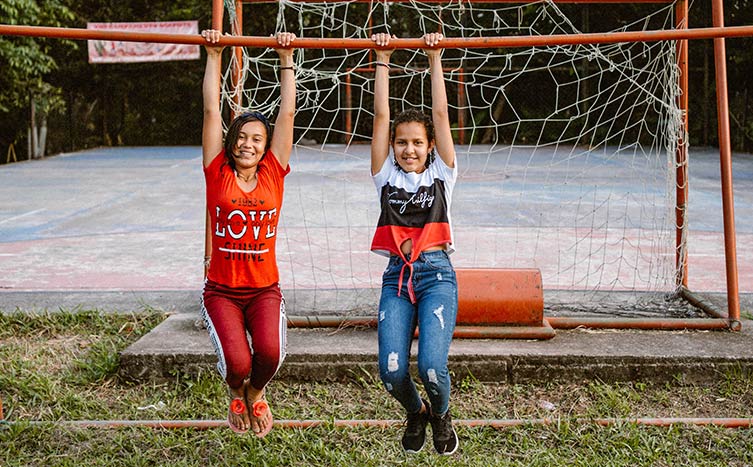 Zwei Mädchen turnen an einer Stange vor einem Fußballplatz