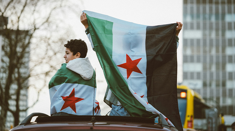 Stadtbild in Syrien. Zwei Personen sitzen eingehüllt in den Revolutionsflaggen auf einer Mauer.