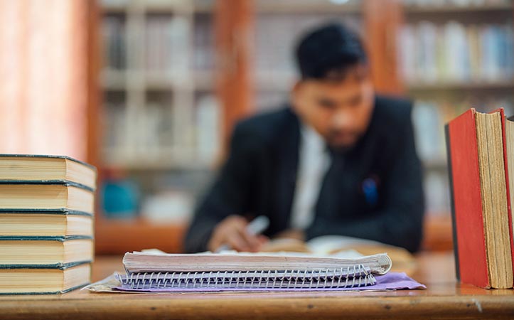 Im Vordergrund sind Bücher und Schreibutensilien zu sehen. Im Hintergrund sitzt ein Mann an einem Schreibtisch
