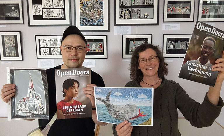 Ein Mann und eine Frau stehen vor einer Wand mit Bildern und halten Bilder und Open Doors Hefte in der Hand