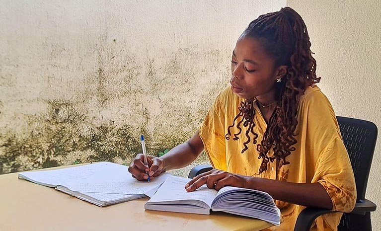 Eine afrikanische Frau macht sich Notizen zu einem aufgeschlagenem Buch. Sie trägt ein gelbes Oberteil.