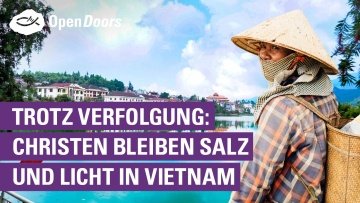 Eine Person mit einem vietnamesischen Hut auf dem Kopf steht vor blauem Gewässer und im Hintergrund sind einige große Häuser zu sehen
