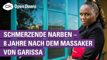 Eine Frau im schwarzen Oberteil mit einer roten Tasche im Arm steht an einem Fenster mit türkisfarbenem Rahmen