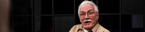 Ein älterer Mann mit Schnurrbart, Brille und beigem Hemd spricht enthusiastisch in einem Interview