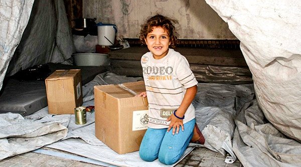 Syrisches Mädchen in einem Planenzelt mit einer Kiste mit Lebensmitteln von der Kirche