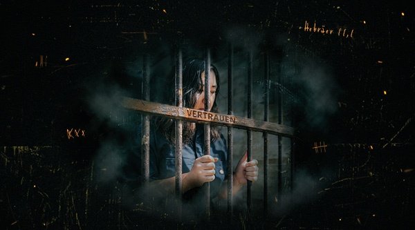 Eine Frau im Gefängnis hält sich an den Gitterstäben fest und schaut traurig nach unten. Auf den Gitterstäben ist das Wort Vertrauen eingraviert.