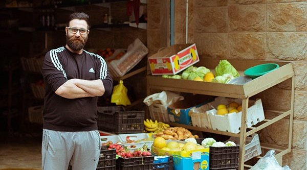 Ein Mann steht mit verschränkten Armen vor mehreren Regalen mit Obst und Gemüse