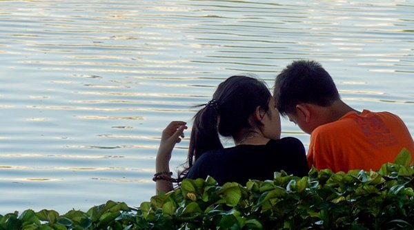 Symbolbild: Ein junges Paar sitzt hinter einer grünen Hecke am stillen Gewässer