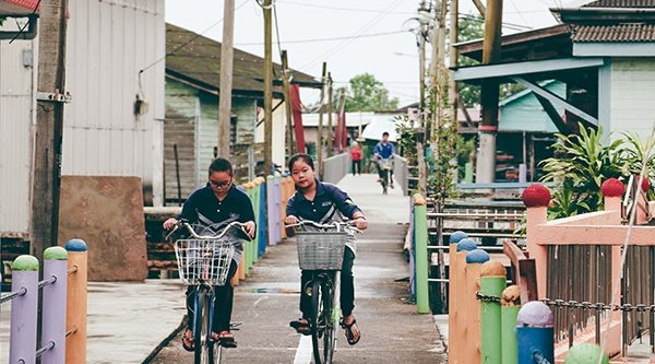 Zwei malaysische Mädchen fahren auf dem Fahrrad frontal auf die Kamera zu und hinter ihnen sieht man die von Häusern umgebene Gasse