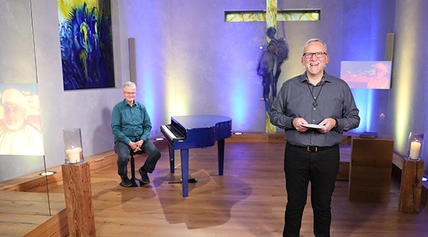 Ein Mann spricht zu den Zuhörern, hinter ihm ein Raum, der ähnlich wie eine Kirche aussieht, mit einem Kreuz als Fenster ganz vorne, Holzfußboden, hohen bunten Glasfenstern und ein Mann sitzt an einem blauen Flügel