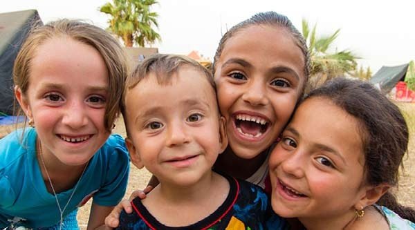 Vier lachende Kinder beugen sich runter zur Kamera, im Hintergrund ist trockenes Gras und Zelte