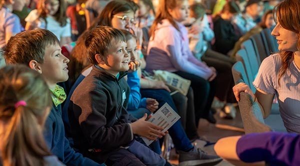 Ein paar Kinder schauen gebannt und mit leuchtenden Augen auf Manu, die vor ihnen sitzt und spricht