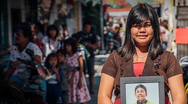 Eine junge indonesische Frau hält das Bild von einem indonesischen Jungen im Bilderrahmen vor sich, im Hintergrund eine Straßenszene