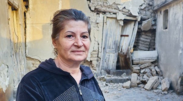 Eine syrische Frau steht vor dem Hintergrund eines in sich gefallenes Betonhauses mit Sonnenlicht an der Hauswand