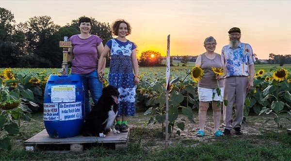 Ein älteres Ehepaar, deren Tochter und Enkelin und ein Hund stehen vor einem Sonnenblumenfeld, links und rechts eine Kassentonne mit Aufschrift