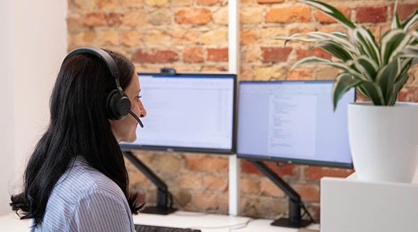 Eine lächelnde Frau mit schwarzen Haaren sitzt mit Headset auf dem Kopf vor zwei Bildschirmen und einer Ziegelwand am Schreibtisch