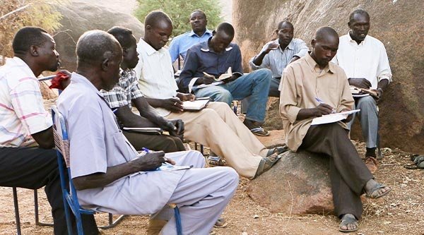 Afrikanische Männer sitzen zwischen Felsen auf Stühlen und schreiben etwas auf Blöcke. 