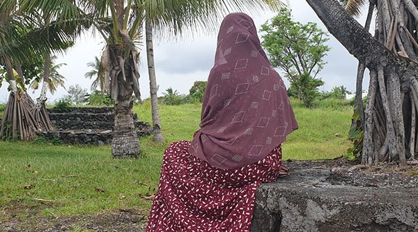 Eine Frau in ein Tuch gehüllt sitzt auf einem Stein und blickt auf ein Gewässer