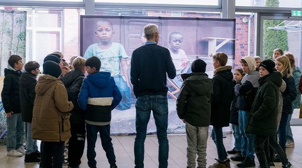 Ein Mann und eine Gruppe Kinder stehen in einem Halbkreis vor einem Bild.